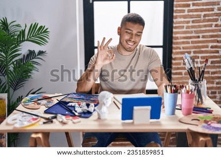Young hispanic painter man doing online call with tablet doing ok sign with fingers, smiling friendly gesturing excellent symbol 