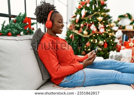 African american woman listening to music sitting on sofa by christmas tree at home Royalty-Free Stock Photo #2351862817