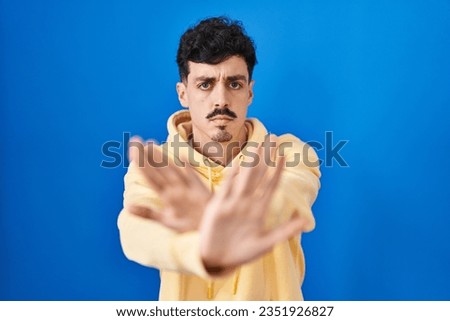 Hispanic man standing over blue background rejection expression crossing arms and palms doing negative sign, angry face 
