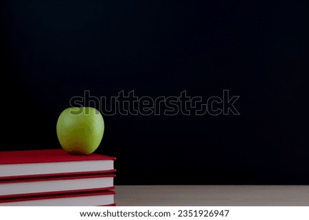 A stack of books with a green apple on the desk on a black background of the blackboard with a place for the inscription.