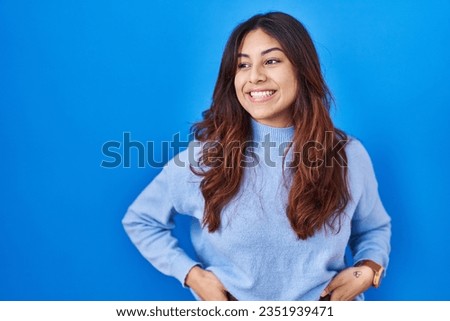 Hispanic young woman standing over blue background looking away to side with smile on face, natural expression. laughing confident. 