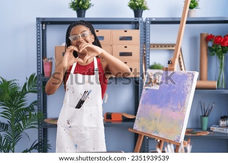 Young african american with braids at art studio painting on canvas smiling in love showing heart symbol and shape with hands. romantic concept. 