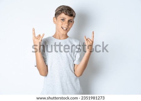Beautiful kid boy wearing grey casual t-shirt makes rock n roll sign looks self confident and cheerful enjoys cool music at party. Body language concept. Royalty-Free Stock Photo #2351981273