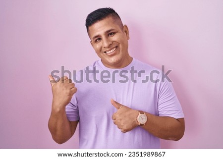 Young hispanic man standing over pink background pointing to the back behind with hand and thumbs up, smiling confident 