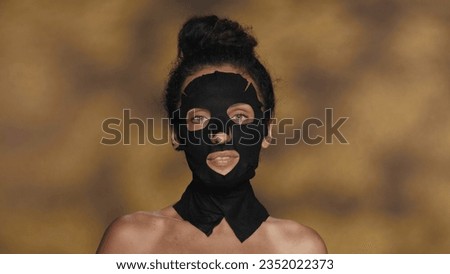 A woman in a black cosmetic sheet mask and with a bun on her head. Seminude woman performing cosmetic procedures in the studio on a yellow background with highlights.