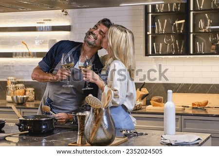 Loving wife kissing husband on cheek while cooking together at home and standing with glasses of white wine at stove Royalty-Free Stock Photo #2352024505