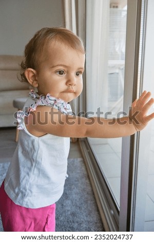 Side view of curious toddler gazing out window with interest and exploring surroundings of house in living room at home
