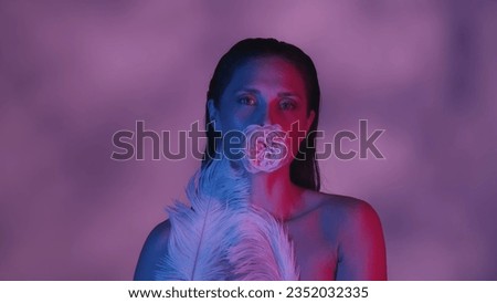 Close up shot of a young woman's face and body. She's confidently looking at the camera with a rose in her mouth and feathers covering part of her body in neon color scheme.