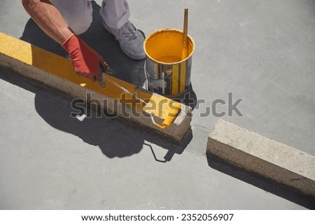 Unrecognizable professional painter at work. Young man uses a paint roller to apply yellow paint for road marking on a parking lot.	