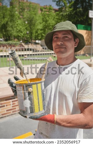 Professional painter at work. Young man holds a bucket of yellow acrylic paint for road marking on asphalt of a parking lot.	
