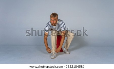 Young handsome blond man in casual t-shirt and beige pants on white background lacing his shoes looking to the side. casual concept Royalty-Free Stock Photo #2352104581