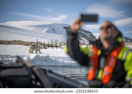 A man on a boat  is taking a selfie picture with a polar bear  walking on a snow in the back.