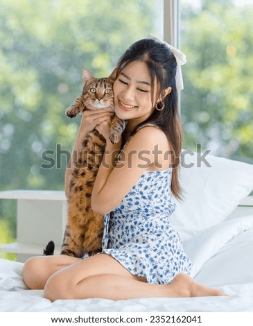 Asian young pretty cheerful female  girl in dress sitting on white clean sheet blanket bed smiling playing hugging holding cute domestic short hair pet cat companion friend in bedroom.