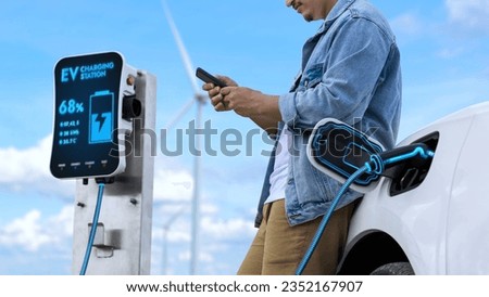 Asian man using smartphone while electric car recharging energy from EV charger from charging station at wind turbine farm. Alternative sustainable and clean energy utilization concept. Peruse