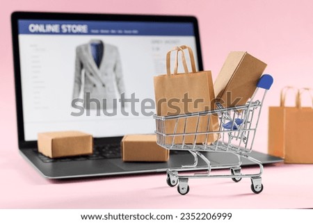 Online store. Mini shopping cart, purchases and laptop on pink background, selective focus