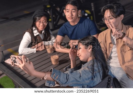 The lady in a denim jacket is holding up a phone to take a picture of herself and with her friends, two of them are ladies and two guys. Motorcycle, parking lot and rail in the background