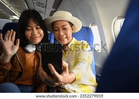 Smiling middle aged woman and daughter sitting in passenger airplane and taking picture, waiting for airplane landing
