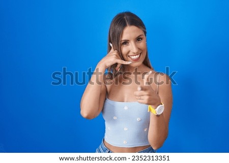 Young woman standing over blue background smiling doing talking on the telephone gesture and pointing to you. call me. 