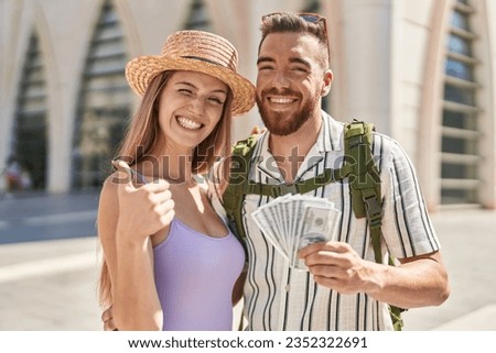 Young tourist couple holding dollars banknotes smiling happy and positive, thumb up doing excellent and approval sign 