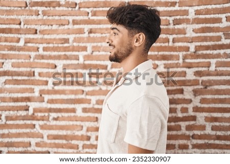 Arab man with beard standing over bricks wall background looking to side, relax profile pose with natural face and confident smile. 