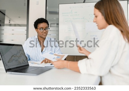 Two businesswomen sitting at the desk and discussing. Diverse female office employees brainstorming in meeting room. HR manager interviewing candidate for the position Royalty-Free Stock Photo #2352333563