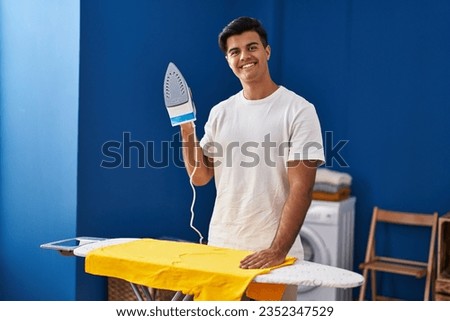 Young hispanic man smiling confident holding iron machine at laundry room