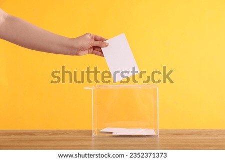 Woman putting her vote into ballot box on wooden table against orange background, closeup Royalty-Free Stock Photo #2352371373