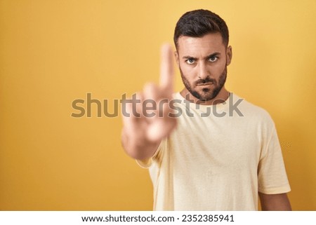 Handsome hispanic man standing over yellow background pointing with finger up and angry expression, showing no gesture 