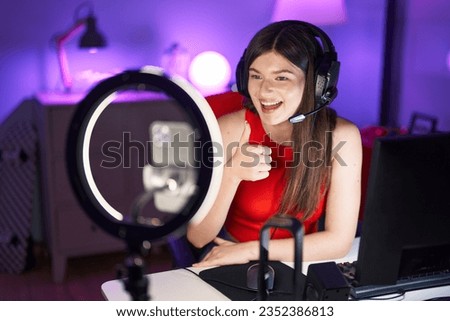 Young caucasian woman playing video games recording with smartphone smiling happy and positive, thumb up doing excellent and approval sign 