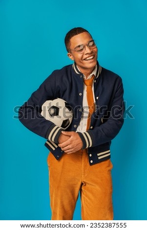 happy african american student in stylish jacket and orange pants holding soccer ball on blue