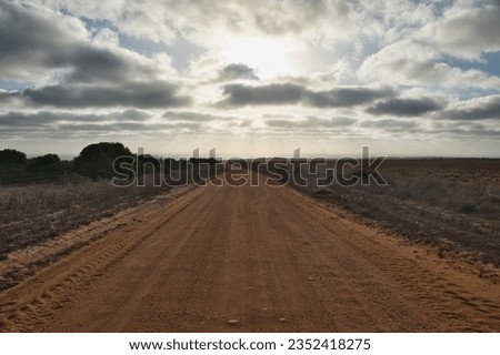 Dirt track leading towards the sunset. Long and wide, empty, red-colored gravel road with sunset in the background. Gravel road and dramatic sky. Western Australia road trip symbol picture.   Royalty-Free Stock Photo #2352418275