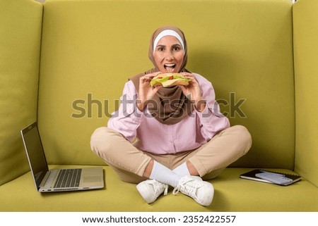 Young Arab girl in a hijab in a cozy workplace has a sandwich after stopping work or learning. Woman holds a burger, bites off healthy food with pleasure. Take a work break for lunch. Green background Royalty-Free Stock Photo #2352422557