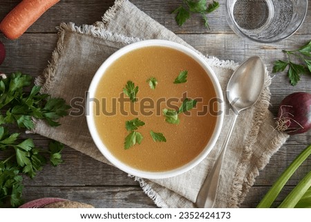 Bone broth made from chicken in a white soup bowl with fresh vegetables on a table, top view