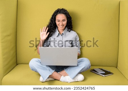 Happy Oriental businesswoman in glasses waving hand at webcam laptop, having meeting in coworking. Hi, greeting and hello, video call, study work and business remotely. Curly girl sits on a green sofa