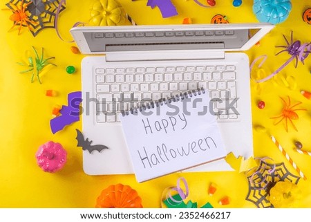 Funny high-colored Halloween bright yellow composition. White laptop with typing hands,  Halloween decor colorful holiday accessories - spiders, cobwebs, pumpkins, bats, top view flat lay copy space