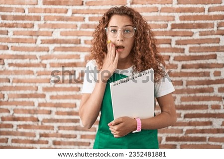Young caucasian woman holding art notebook afraid and shocked, surprise and amazed expression with hands on face 
