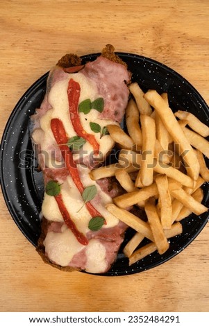 Top view of milanesa dish, fried breaded steak, with mozzarella cheese, ham, oregano, grilled red bell pepper and fried potatoes, on the wooden table. 
