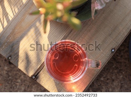 Transparent teacup with happy smiley pattern, filled with pink drink. Top-down shot on wooden background, surrounded by plants in nature. Good vibes.





 Royalty-Free Stock Photo #2352492733