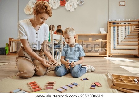 montessori school, female teacher sitting near blonde boy and showing wooden toys, educational game Royalty-Free Stock Photo #2352500611
