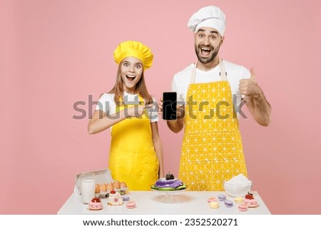 Teen fun girl dad chef cook confectioner baker in yellow apron cap at table show mobile phone blank screen workspace thumb up isolated on pink background Mousse cake food workshop master class process