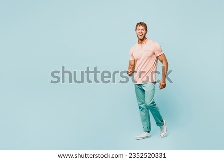 Full body sideways smiling happy fun young caucasian blond man he wear pink t-shirt casual clothes look camera walk go isolated on pastel plain light blue background studio portrait. Lifestyle concept