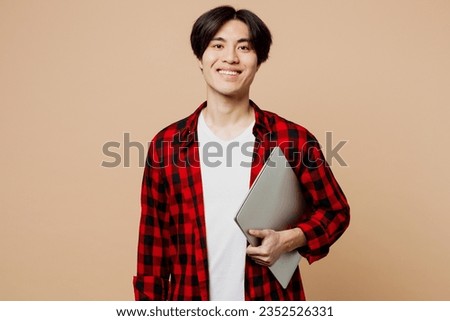 Confident happy smiling smart young IT man of Asian ethnicity he wear red shirt casual clothes hold closed laptop pc computer isolated on plain pastel light beige background studio. Lifestyle concept Royalty-Free Stock Photo #2352526331