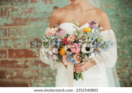 A bride holding a beautiful bouquet of colorful flowers wearing a unique, off shoulder bridal gown in front of a patina teal blue exposed brick wall photographed on film
