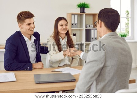 Happy cheerful business partners or young family couple shake hands with loan broker, estate agent or bank manager as they sign contract agreement and make successful deal during meeting in office