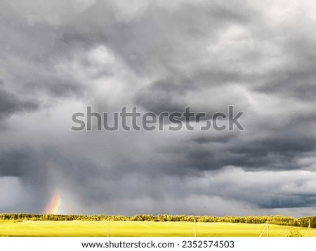 Nature landscape with gloomy stormy sky, gloomy dramatic low gray clouds and a piece of colored rainbow over a field with bright green and yellow grass on spring, autumn or summer day or evening