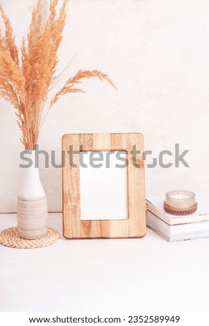 Mockup wooden picture frame, books, aroma candle and pampas grass in vase on white table. Copy space.