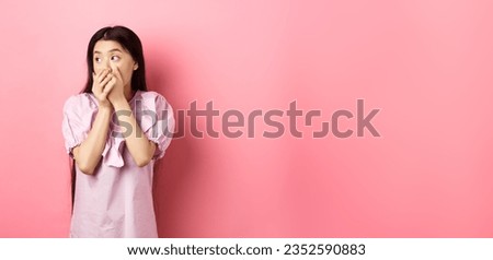 Shocked and startled asian woman looking aside at logo, covering mouth with hands speechless, standing in dress on pink background. Royalty-Free Stock Photo #2352590883