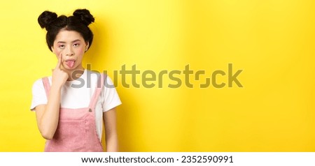 Rude asian girl stretch eyelid and showing tongue, mocking someone, standing on yellow background. Royalty-Free Stock Photo #2352590991