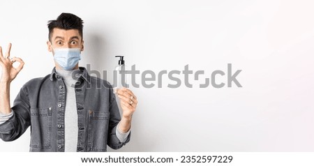 Healthy people and covid-19 concept. Excited man in medical mask holding bottle of good hand sanitizer, show okay sign, recommend antiseptic, standing against white background.