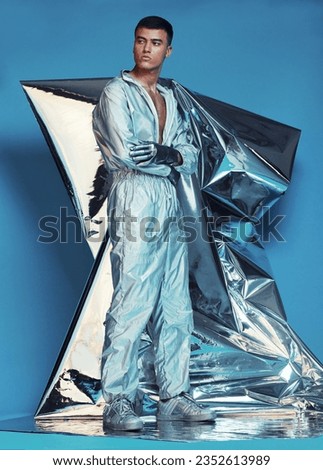Fashion, cyberpunk and silver with a man on a blue background at a photo shoot in a shiny futuristic outfit. Sci fi, creative or art with a young model in studio for holographic or vaporwave clothes